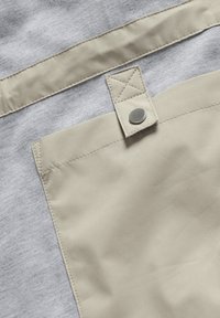 Poche en tissu beige avec un rabat boutonné fixé à un fond en tissu texturé gris clair.