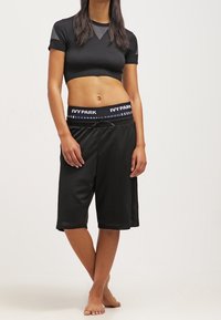 Mujer con top corto negro y pantalones cortos de malla sueltos negros con una cintura de "IVY PARK", de pie descalza frente a un fondo claro y liso.