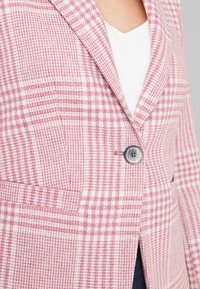 Blazer à carreaux rose et blanc avec des revers en notch, fermeture à un bouton et poches latérales. Texture douce avec une coupe structurée.