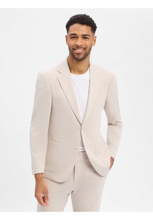 Lächelnder Mann mit kurzen lockigen Haaren, der eine beigefarbene Anzugsjacke und Hose über einem weißen T-Shirt trägt, vor einem schlichten weißen Hintergrund stehend.