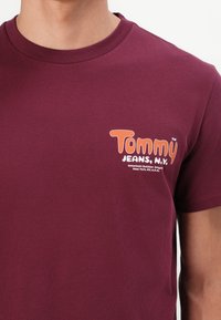 Person, der ein kastanienfarbenes T-Shirt mit kurzen Ärmeln und einem orange-weißen "Tommy Jeans, N.Y."-Logo auf der Brust trägt.