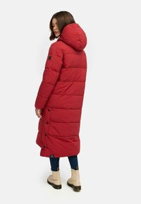 Red padded long coat with a hood, featuring horizontal quilting, side buttons, and a label on the hem. Worn over blue jeans and boots.