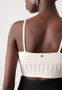 Helle beige Bralette mit verstellbaren Trägern, strukturiertem Strickdesign, komplizierten Mustern und einem kleinen Logodetail unter dem Rückenausschnitt.