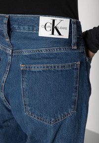 Primo piano della tasca posteriore di un paio di jeans in denim blu con una patch bianca del logo Calvin Klein Jeans sopra la tasca, indossati con un top nero.