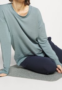 Persona seduta su un tappetino grigio, indossando una maglietta lunga a maniche larghe blu-verde e pantaloni scuri blu navy, con le mani appoggiate sul pavimento e sul ginocchio.