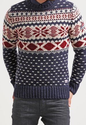 Marineblauer Strickpullover mit roten und weißen nordischen Mustern und geripptem Saum, getragen über einem dunklen Hemd mit Kragen bei einer Person mit den Händen in den Taschen.