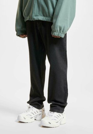 Zwarte spijkerbroek gecombineerd met een groene lichte jas, afgemaakt met witte sneakers met uitsparingen voor ventilatie.