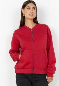 Felpa rossa con zip, polsini e orlo a coste, con due tasche frontali e scollo arrotondato. Realizzata in tessuto morbido e leggermente strutturato.