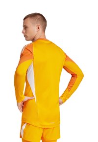Maglietta atletica arancione brillante e gialla con maniche lunghe, dettagli in rete ai lati e design aderente. Gli shorts sono abbinati nel colore.