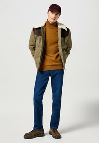 Brun kordflodjacka med krämfärgad faux shearling-krage, orange tröja, blå jeans och bruna skor. Enkelt och funktionellt design.