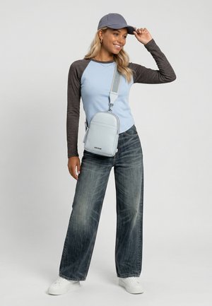 Woman in light blue and dark gray long-sleeve shirt, wide-leg jeans, gray cap, white sneakers, and light blue crossbody bag smiling and posing.