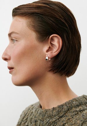 Profilul unei persoane cu păr scurt și castaniu, purtând cercei argintii în formă de cerc și un pulover texturat gri-verzui, pe un fundal simplu.