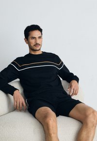 Svart genser med brune og hvite horisontale striper, laget av mykt stoff. Kombinert med matchende svarte shorts, sitter på en lys sofa.