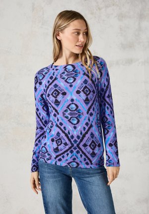 Pull en tricot violet présentant un motif en losanges dans des nuances de bleu et de noir, avec des manches longues et un col rond, associé à un jean.