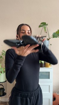 Pantofi sport negri cu un design elegant, finisaj lucios și talpă texturată. Ținuți de o persoană pe un fundal neutru cu plante.