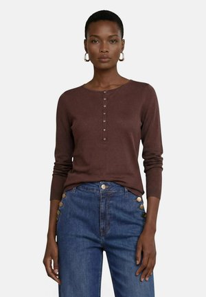 Femme portant un haut marron à manches longues boutonné et un jean bleu avec de gros boutons sur les poches, debout devant un fond uni.