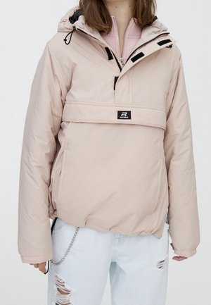 Osoba nosząca jasną beżową puchową kurtkę z kapturem z przednią klapką i jasnoniebieskie porwane dżinsy.