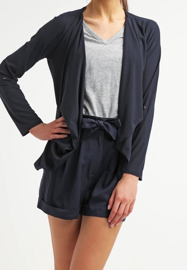 Marinblå öppen cardigan med långa ärmar över en grå t-shirt med V-ringning, i kombination med högmidjade marinblå shorts med en rosettdetalj.