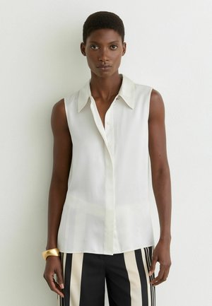 Femme aux cheveux courts portant un chemisier blanc sans manches boutonné et un pantalon noir et blanc à rayures verticales, debout contre un mur blanc.