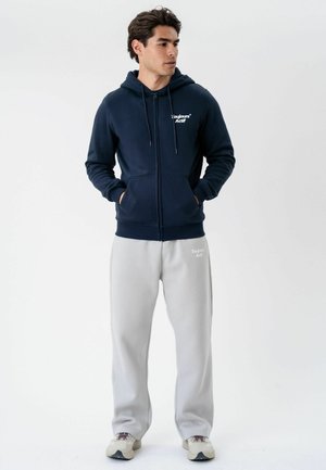 Young man standing with hands in pockets wearing a navy zip-up hoodie and light gray sweatpants with white sneakers.