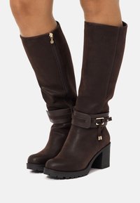Botas de imitación de cuero marrón hasta la rodilla con un tacón cuadrado grueso. Presentan una cremallera lateral y una correa decorativa alrededor del tobillo con una hebilla metálica.