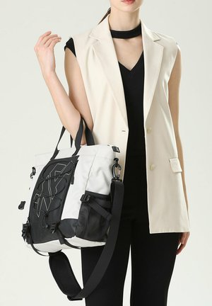 Grand sac fourre-tout blanc et noir, fabriqué en tissu durable, avec une bandoulière réglable, des poches latérales en mesh et un design de logo bien en évidence.