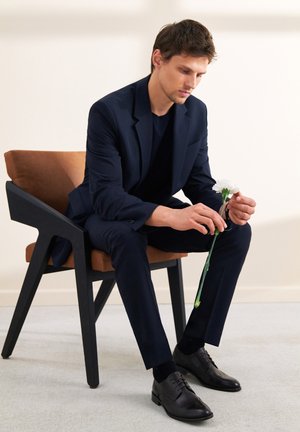 Granatowy garnitur z pasującą koszulą, siedzący na czarnej drewnianej krześle z brązowym poduszką. Trzymając białą goździk, w czarnych butach wyjściowych.