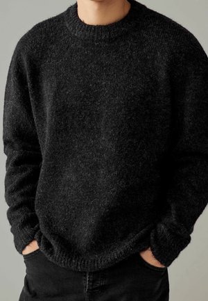 Personne portant un pull noir duveteux à col rond, les mains dans les poches d’un pantalon noir, debout devant un fond gris uni.