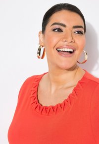 Top rojo con un diseño de escote con volantes. Grandes pendientes de aro dorados y un maquillaje suave y ligero completan el look.