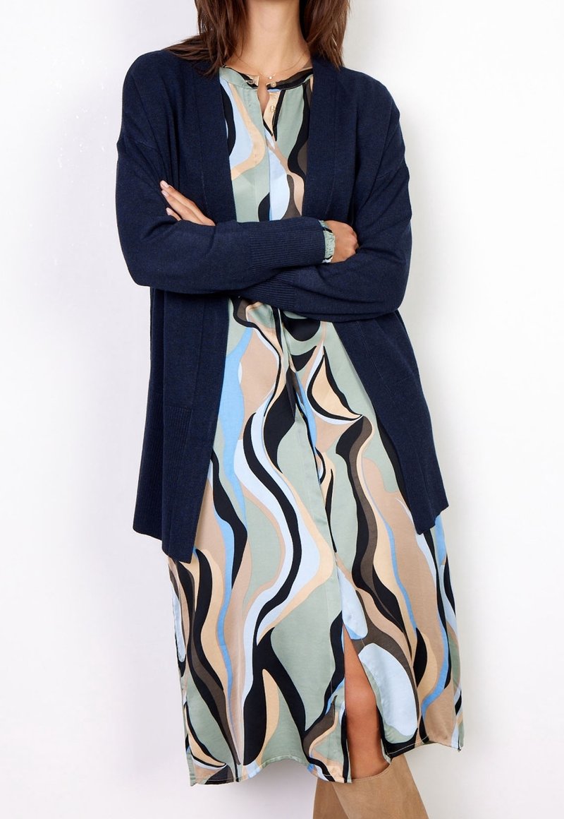 Femme portant une robe fluide à motif abstrait multicolore, avec un cardigan bleu marine ouvert devant et des bottes beige, bras croisés.