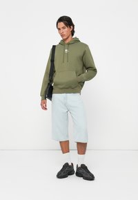 Olijfgroen hoodie met een voorzak en een koord, gecombineerd met lichtblauwe denimshorts tot halverwege de dij. Zwarte sneakers en witte sokken.