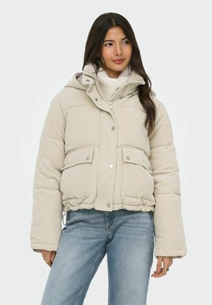 Femme portant une veste en velours côtelé matelassée beige par-dessus un pull à col roulé en tricot blanc et un jean bleu, debout devant un fond uni.
