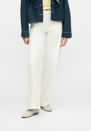 Person iført hvide, højtaljede bukser med vide ben, beige flade sko, gul top og kort, mørk denimjakke mod en ensfarvet baggrund.