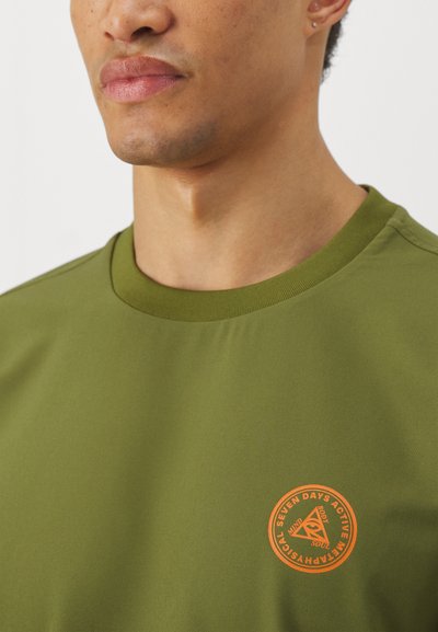 Olive green t-shirt with a round neckline and short sleeves. Features a circular orange logo on the chest, smooth fabric texture.