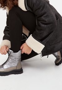 Botines plateados metálicos de tobillo con un diseño acolchado, que cuentan con un cierre lateral y una suela texturizada, combinados con leggings negros y un abrigo negro.