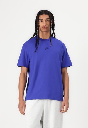 Purpurinė medvilninė marškinėliai su trumpomis rankovėmis, apvaliu kaklu ir mažuoju juodu Nike logotipu ant krūtinės. Modelis dėvi šviesiai pilkas sportines kelnes.