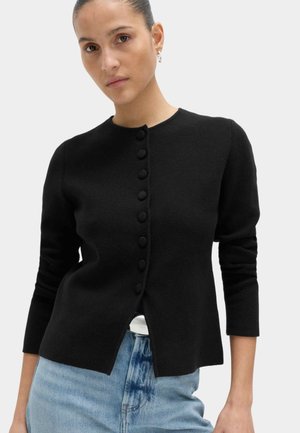 Frau mit nach hinten gekämmtem Haar, die kleine Creolenohrringe, eine schwarze Strickjacke mit Knöpfen und hellblaue Jeans vor einem schlichten Hintergrund trägt.