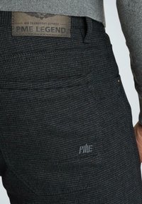 Czarne spodnie o fakturowanej powierzchni z subtelnym wzorem, z skórzaną metką oznaczoną "PME LEGEND" oraz małym logo na tylnej kieszeni.
