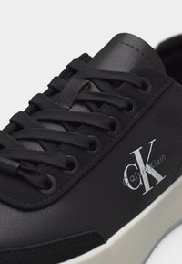 Μαύρο αθλητικό παπούτσι χαμηλού προφίλ με μαύρα κορδόνια, δερμάτινη υφή, λευκή σόλα και λευκό λογότυπο "CK Calvin Klein" στο πλευρό.