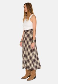 Jupe maxi à carreaux dans les tons marron, écru et beige, avec taille haute et tissu léger. Associée à des bottes de cow-boy beiges et un débardeur blanc.