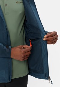 Giacca blu con zip, dotata di un interno in rete e una tasca con zip con un pomello arancione brillante, indossata sopra una polo verde.