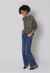 Långärmad topp med leopardmönster i grått och svart, kombinerad med blå utsvängda jeans och svarta klackskor med spetsig tå. Modellen står med händerna i fickorna.