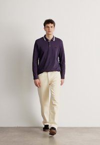 Μακρυμάνικη πόλο μπλούζα σε ναυτικό χρώμα με αντίθεση λευκή τρέσα στον γιακά, συνδυασμένη με παντελόνι σε κρεμ απόχρωση και σκούρα υποδήματα σε απλό φόντο.