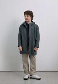 Regnrock i mörkgrönt, vattentätt material, knäppning fram, huva, kombinerad med en mörk tröja och ljusbeige byxor, vita sneakers.