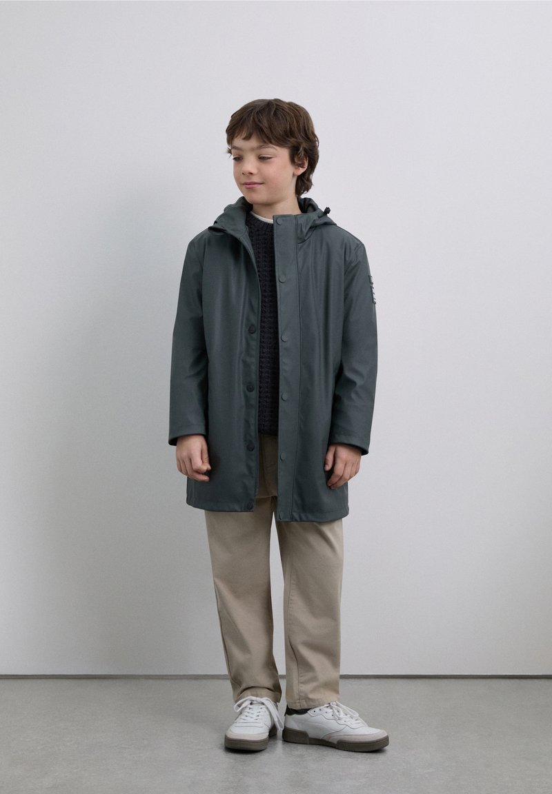 Regnrock i mörkgrönt, vattentätt material, knäppning fram, huva, kombinerad med en mörk tröja och ljusbeige byxor, vita sneakers.