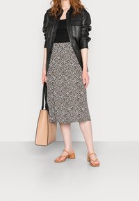 Veste noire en cuir à boutons portée sur un débardeur noir, associée à une jupe blanche mi-longue à imprimé léopard. Sac fourre-tout beige et sandales tan.