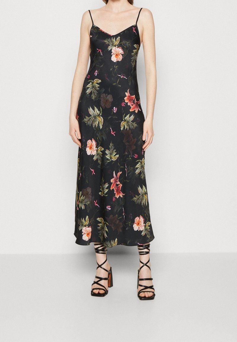 Femme portant une robe nuisette noire à fleurs avec des bretelles fines et des sandales noires à talons hauts et lanières, debout devant un fond uni.