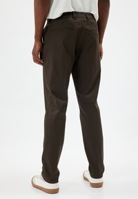 Pantalon marron foncé à coupe droite, doté de deux poches arrière et d'une texture lisse, associé à des baskets blanches.