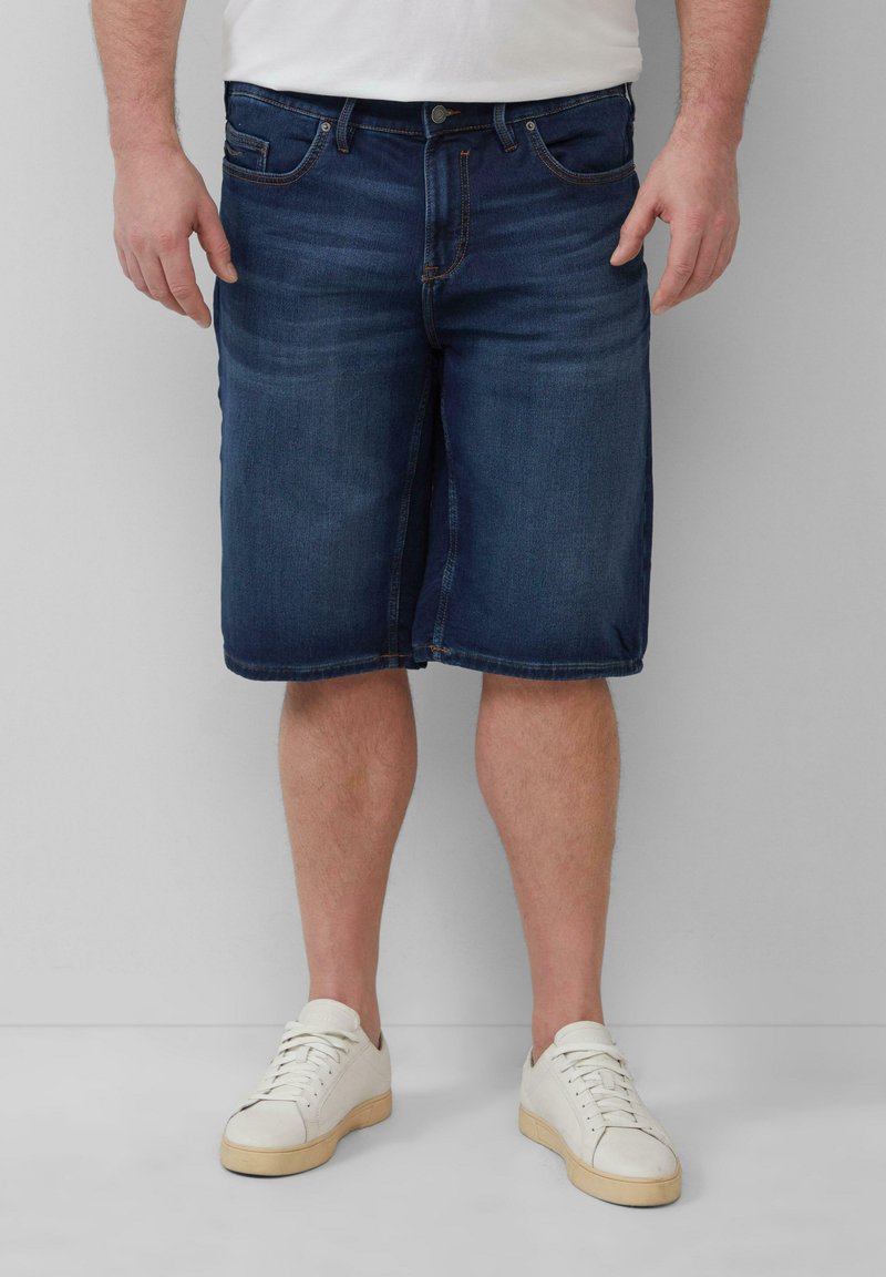 Dunkelblaue Jeansshorts mit geradem Schnitt, fünf Taschen und leichtem Verblassen. Getragen mit weißen Turnschuhen.