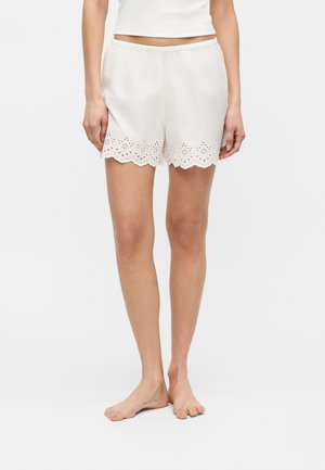 Fehér női rövidnadrág elasztikus derékpánttal és virágos lyukasztott csipke szegéllyel az alján, mezítláb viselve egy fehér, rövid felsővel.